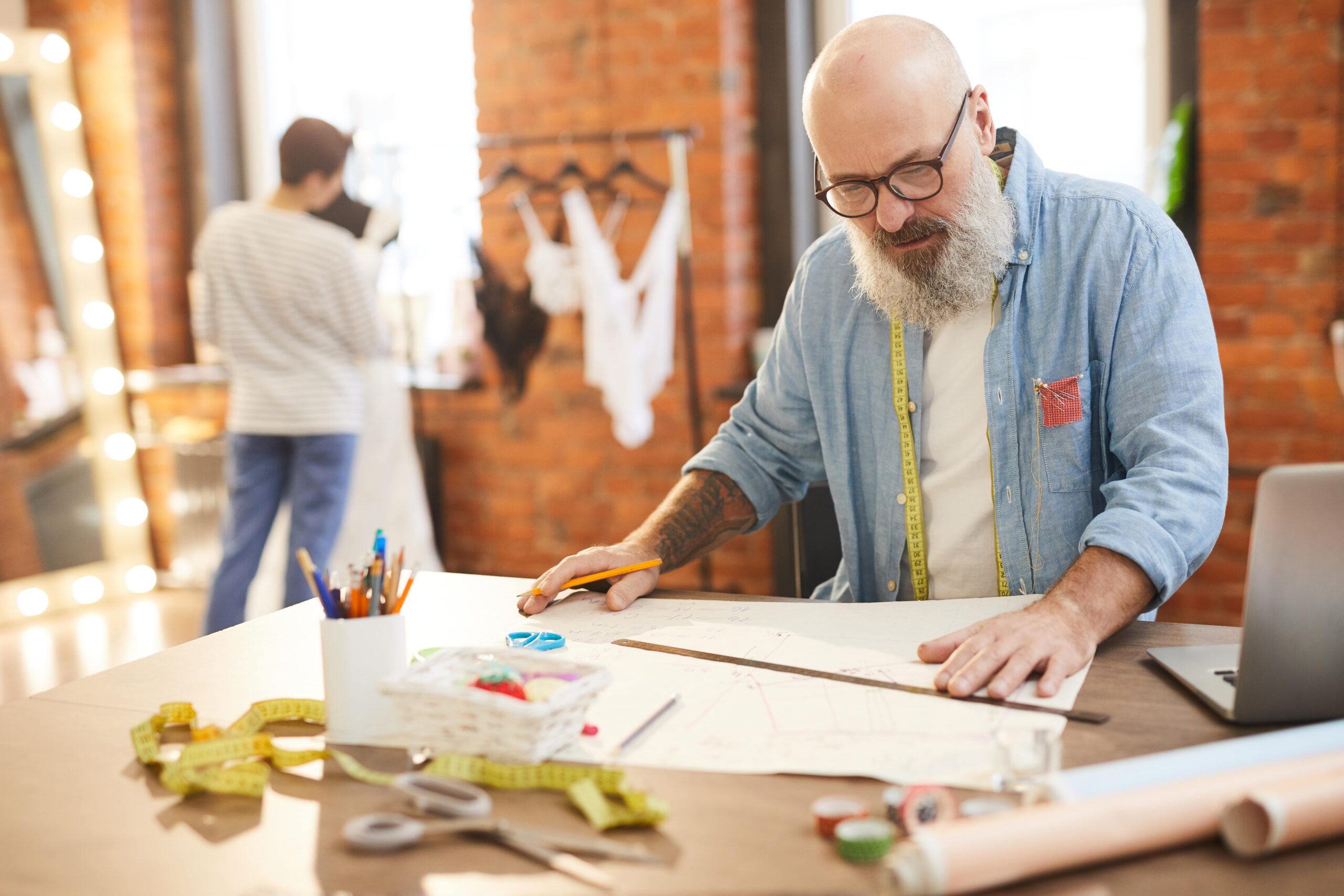 Mature tailor with grey beard uses his laptop to make a new fashion sketch on paper. Internet service by Kinetic Business