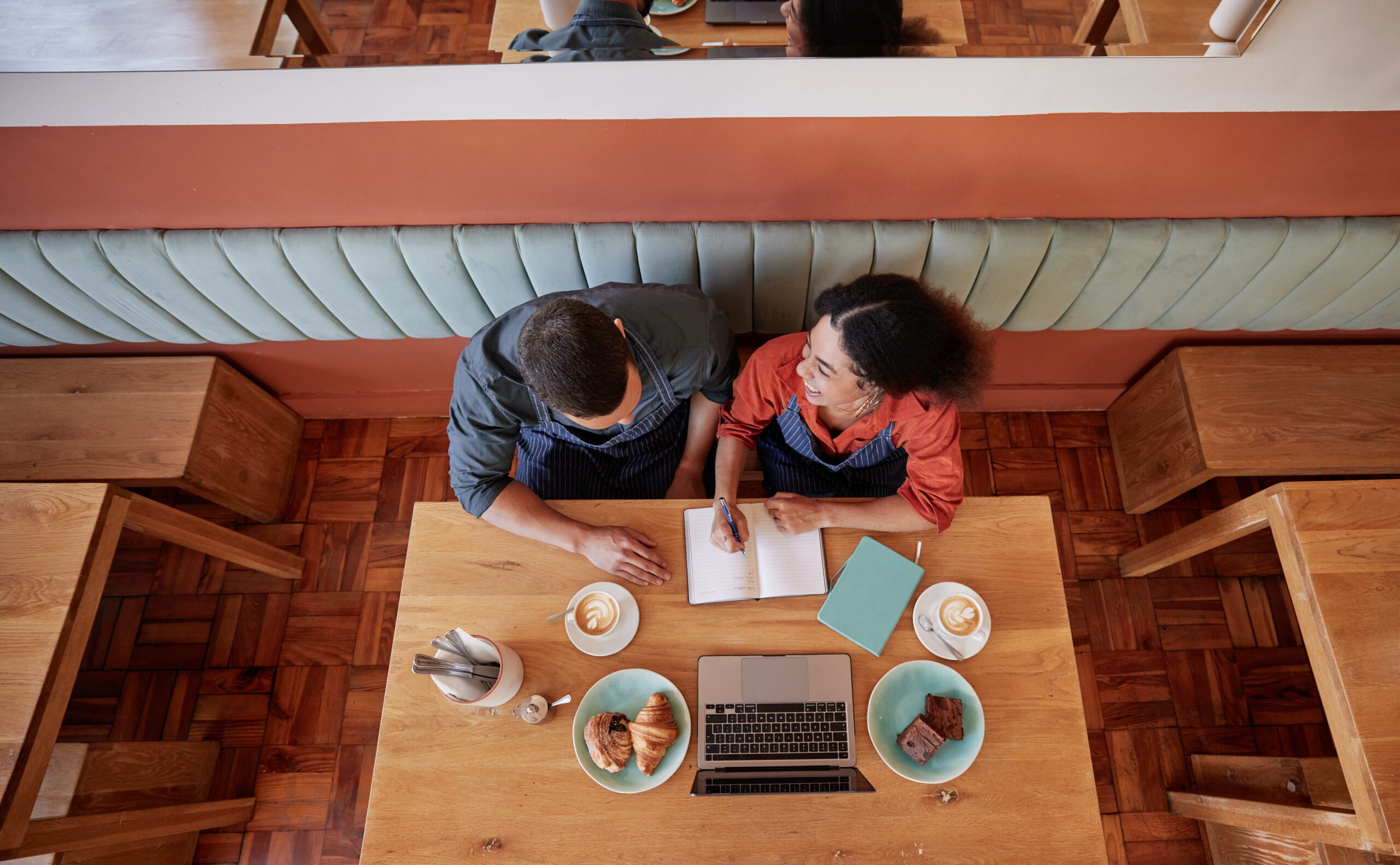 Photo from above of two coffe shop employees wearing aprons planning with the aid of a laptop powered by Kinetic Business