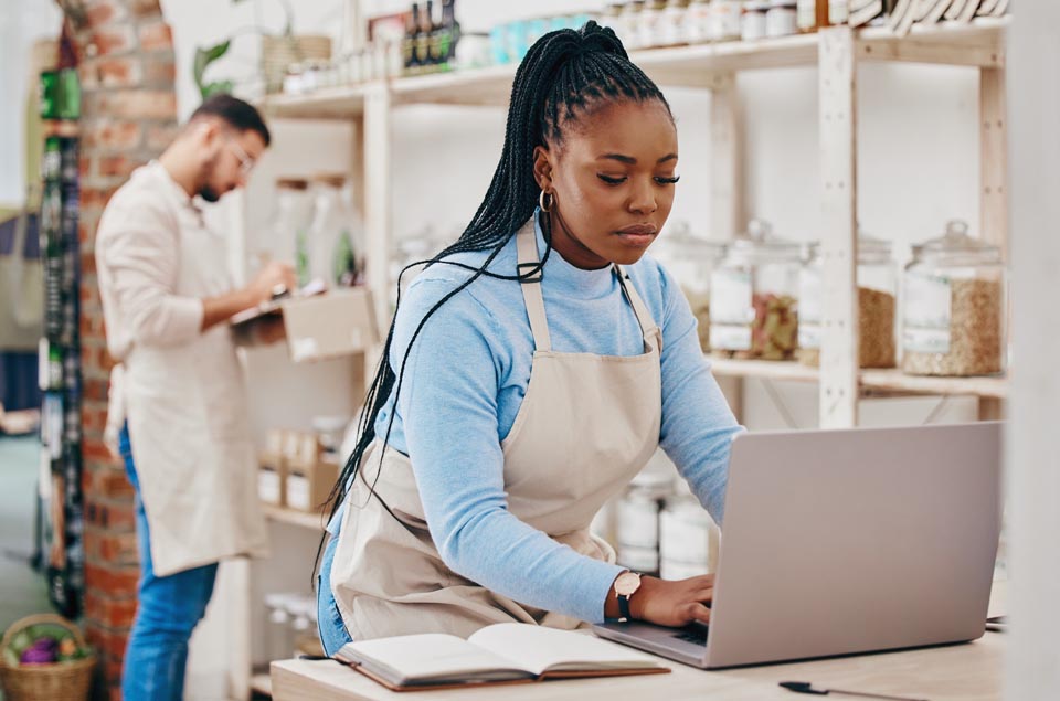 In a small grocery store a woman at the counter uses a laptop to check inventory via a Kinetic Business internet connection