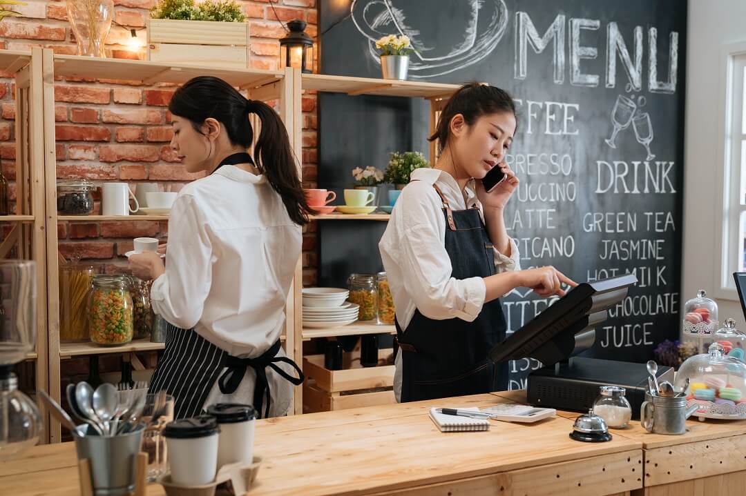 Two female cafe workers using business phone and internet for customer transactions.