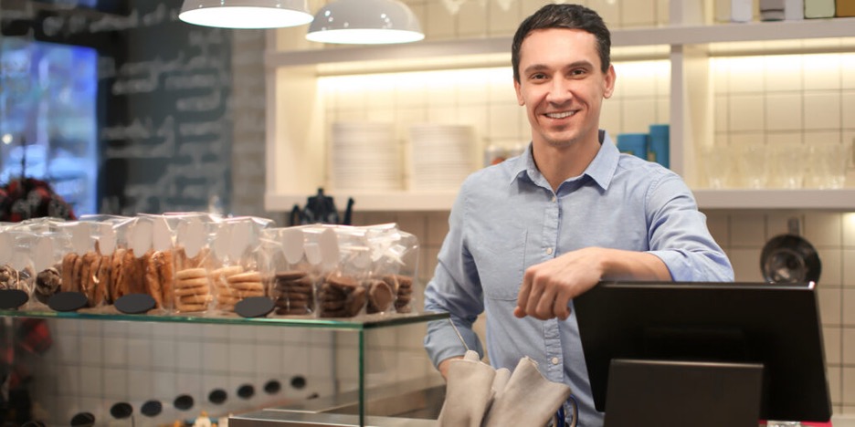 young male baker smiling leaning on computer with baked goods